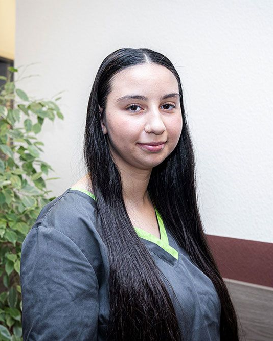 Frau in grauer OP-Kleidung lächelt, langes schwarzes Haar, in der Nähe von Zimmerpflanzen.