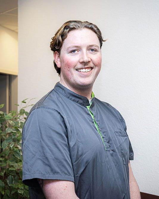 Ein Mann in grauer OP-Kleidung lächelt in einer medizinischen Umgebung und steht neben einer Pflanze.