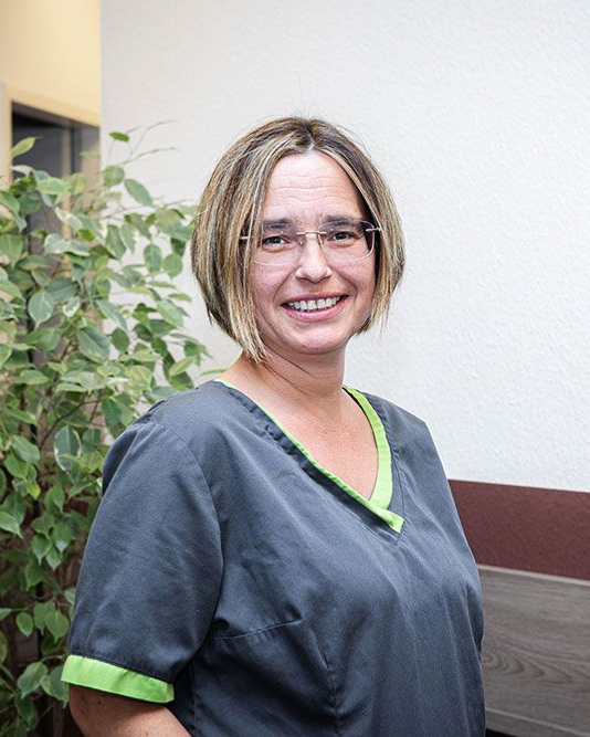 Frau in OP-Kleidung und Brille, lächelnd, vor einer Pflanze und einer Wand, möglicherweise in einer Arztpraxis.