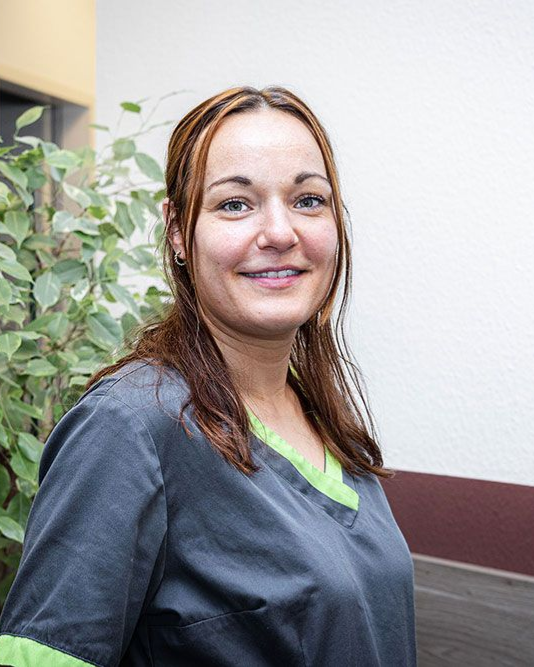 Eine Frau in grauer OP-Kleidung lächelt in einer Klinik, im Hintergrund ist Grün zu sehen.