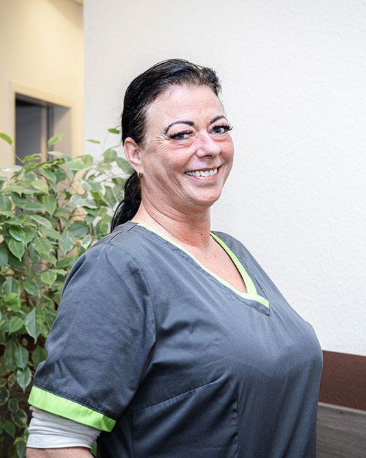 Eine Frau in grauer OP-Kleidung lächelt in einer Arztpraxis mit grünen Zierelementen und einer sichtbaren Pflanze.