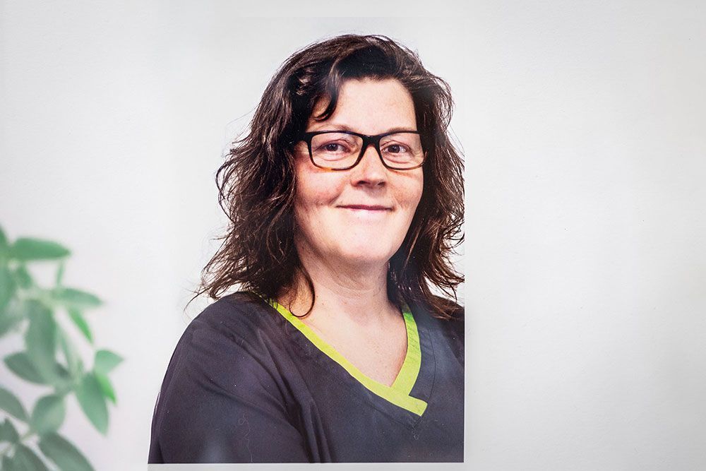 Eine Frau mit dunklen Haaren und Brille lächelt vor einer weißen Wand. Sie trägt ein dunkles Oberteil mit gelbem Saum.