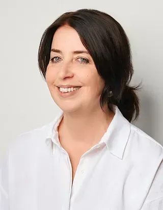 Eine Frau mit dunklen Haaren lächelt und trägt ein weißes Hemd mit Knöpfen.