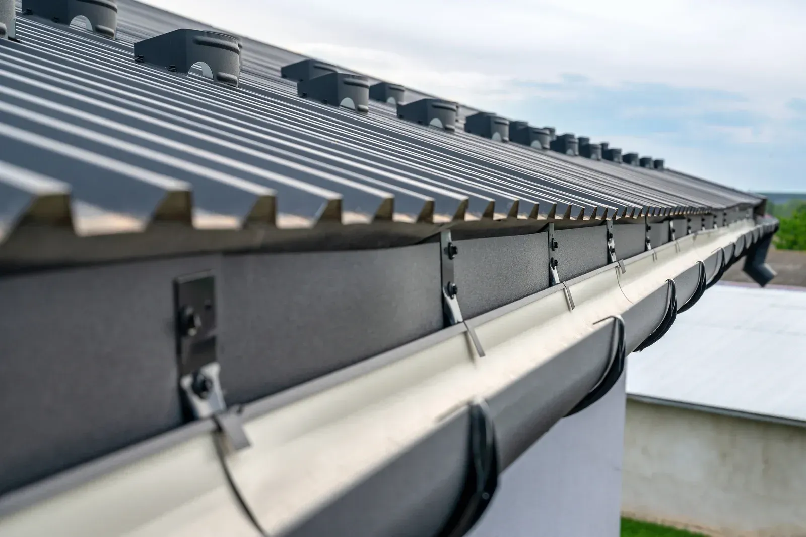 Close-up of a gray metal roof edge with gutters and brackets under a cloudy sky