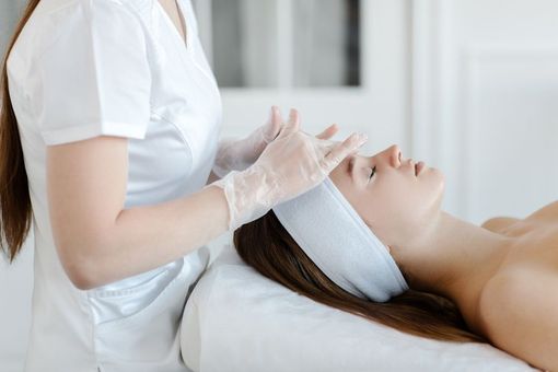Mujer recibiendo un tratamiento facial en un spa. Un técnico con guantes le toca suavemente el rostro. Configuración blanca.