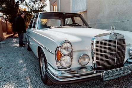 Ein Mann steht neben einem weißen Mercedes Benz, der in einer Einfahrt geparkt ist.
