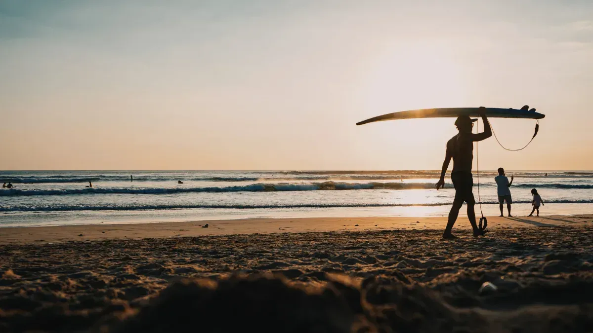 Silueta de una persona que lleva una tabla de surf en la cabeza, caminando por una playa al atardecer.
