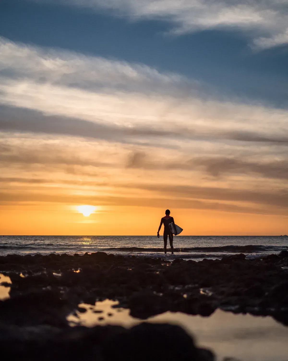 Surfista de pie sobre las rocas al atardecer, sosteniendo una tabla de surf, océano y cielo iluminados en naranja y oro.