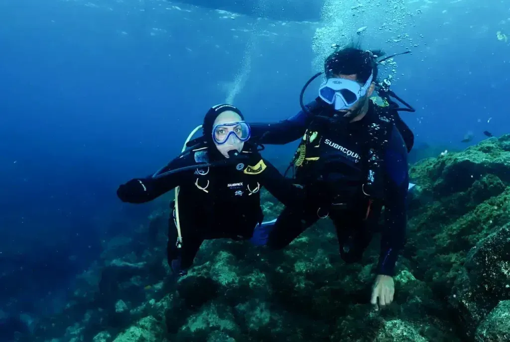 Dos buzos bajo el agua, uno guiando al otro. Agua azul, arrecife de coral.