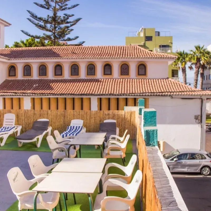 Patio en la azotea con mesas, sillas y tumbonas. Edificio con techo de terracota al fondo.
