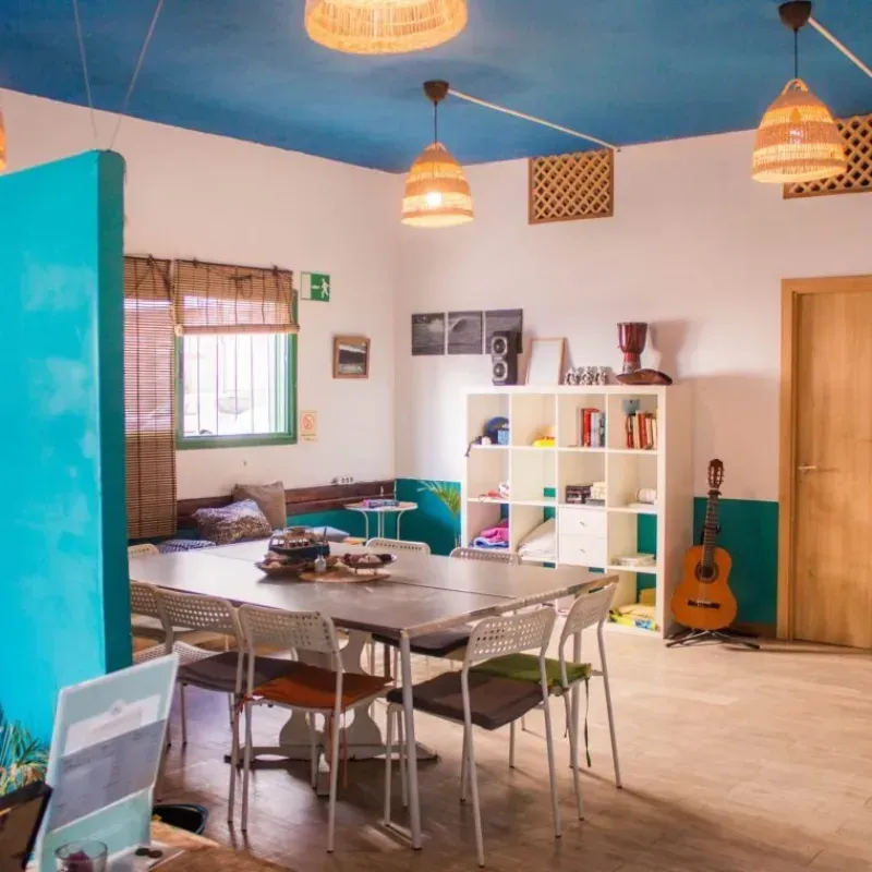 Habitación bien iluminada con mesa, sillas, estanterías y una guitarra. Paredes azules y blancas, lámparas colgantes.