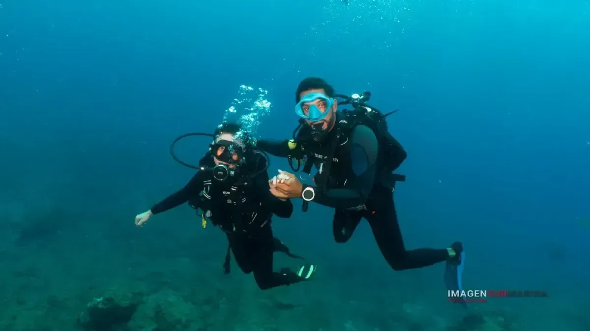 Dos buceadores bajo el agua, uno apuntando, fondo de agua azul.