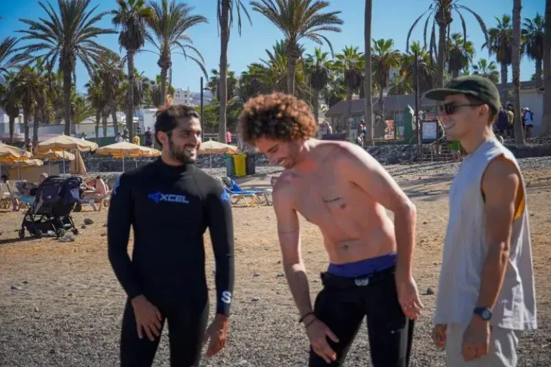 Tres hombres en una playa soleada, uno con traje de neopreno, otro sin camisa y el tercero con gorra.
