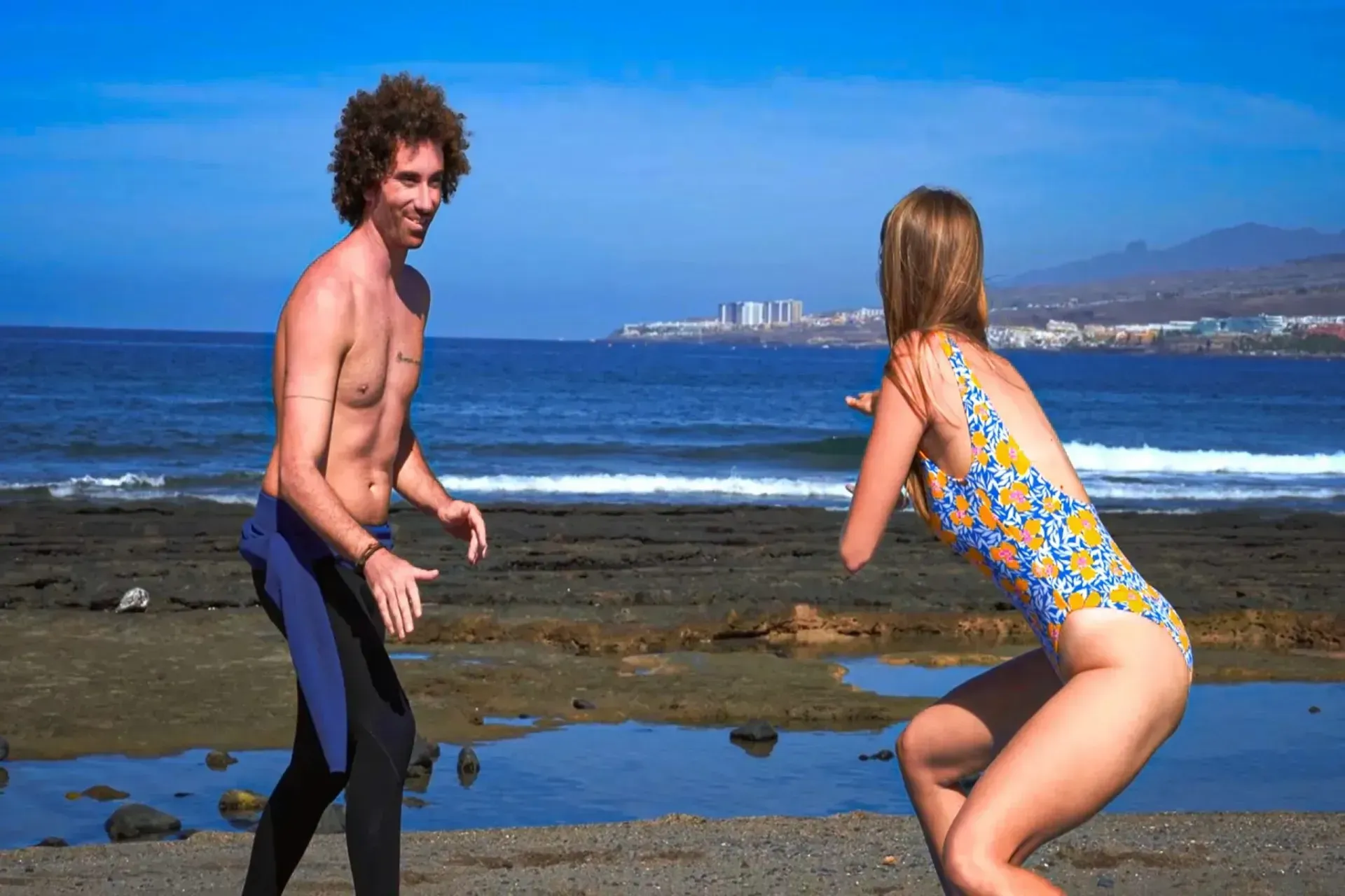 Un hombre y una mujer juegan en la playa; una mujer en traje de baño, un hombre en traje de neopreno.