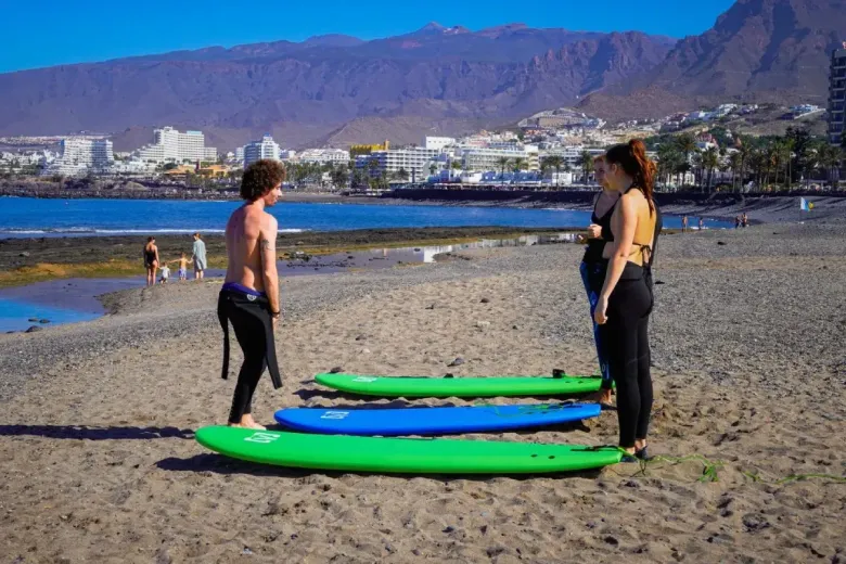 Dos personas en la playa con tablas de surf. Montañas y una ciudad al fondo. Día soleado.