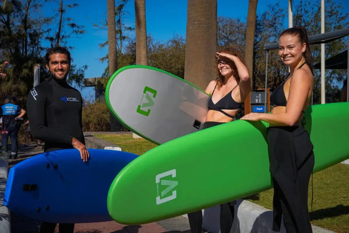 Tres personas con tablas de surf al aire libre. Una lleva traje de neopreno y dos en traje de baño.
