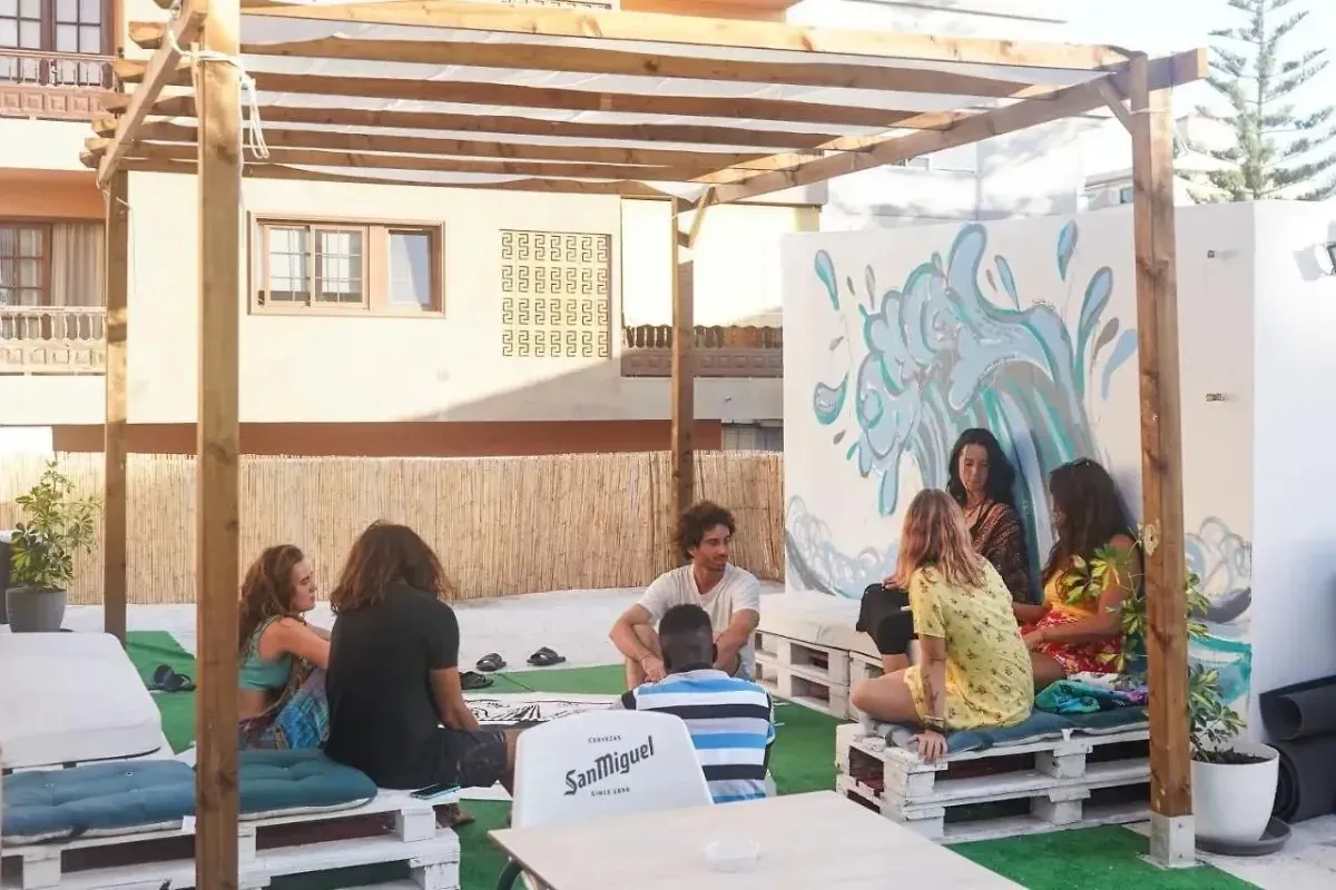 Personas sentadas bajo una pérgola de madera, algunas sobre muebles de palets, cerca de un mural de agua.