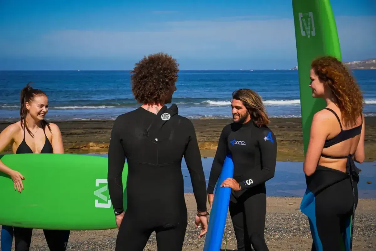 Cuatro personas en trajes de neopreno con tablas de surf en una playa, preparándose para surfear.