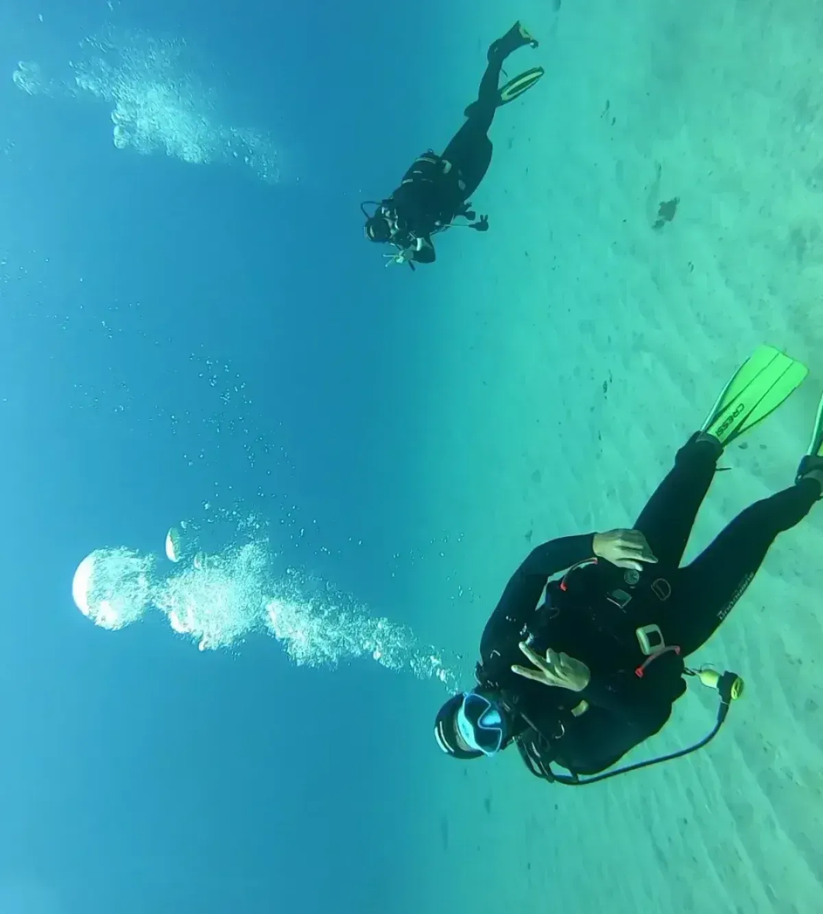 Dos buceadores en el océano, uno haciendo el signo de la paz, con burbujas subiendo. Agua azul, fondo arenoso.