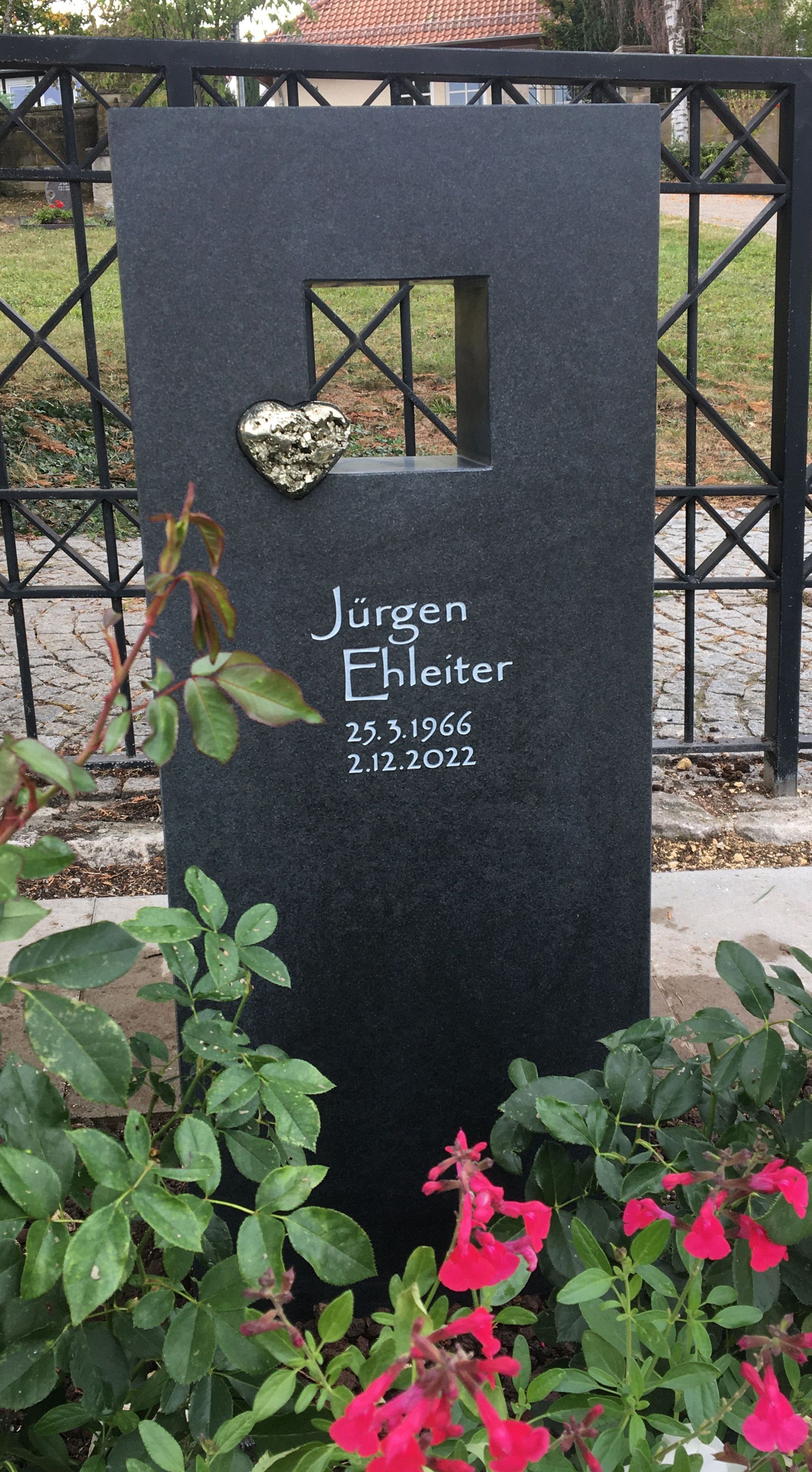 Ein schwarzer Grabstein mit einem Herz darauf ist von Blumen und Pflanzen umgeben.