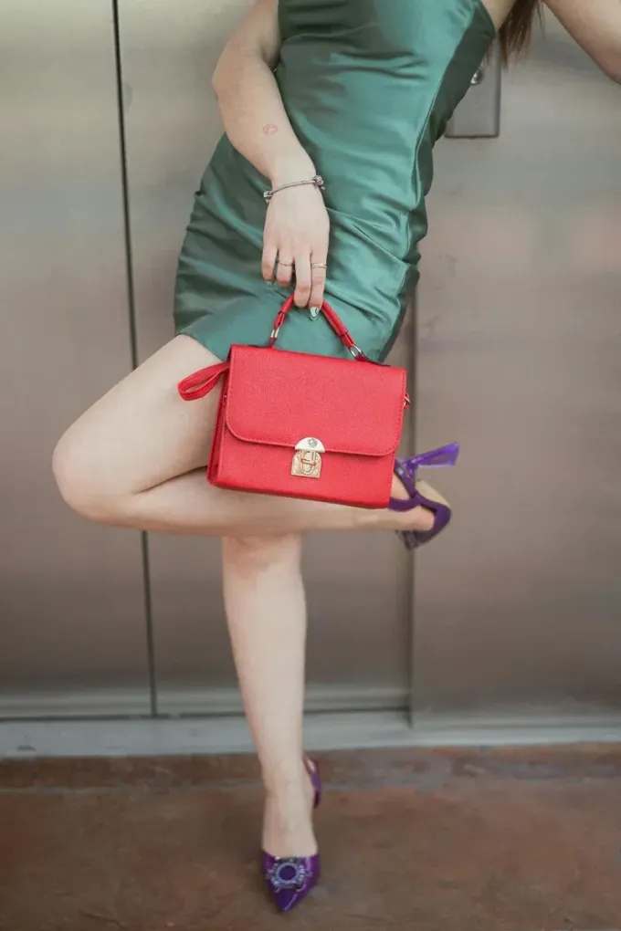 Mujer con vestido verde posa con bolso rojo y tacones morados.