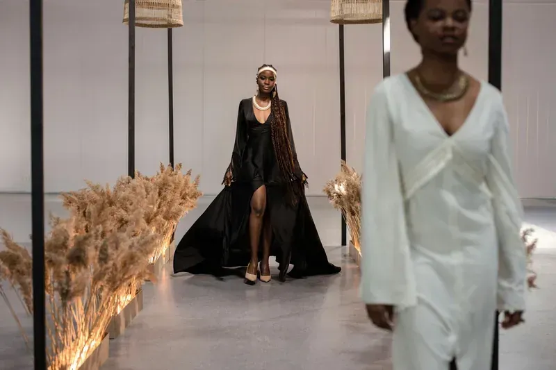 Dos modelos en la pasarela. Una con un vestido negro y la otra con uno blanco. Plantas secas adornan la pasarela.