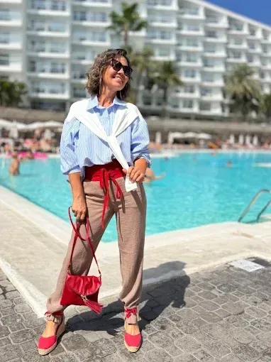 Mujer con camisa azul y pantalones rojos posa junto a una piscina, sosteniendo un bolso rojo y un suéter alrededor de los hombros.