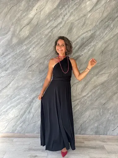 Una mujer con vestido negro de un solo hombro posa sonriendo frente a una pared de mármol gris. Lleva collar y zapatos rojos.