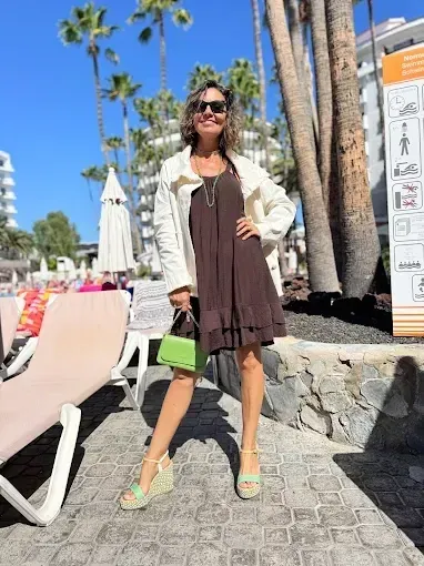 Mujer con vestido marrón y chaqueta color crema, sosteniendo un bolso verde, usando cuñas junto a una piscina.