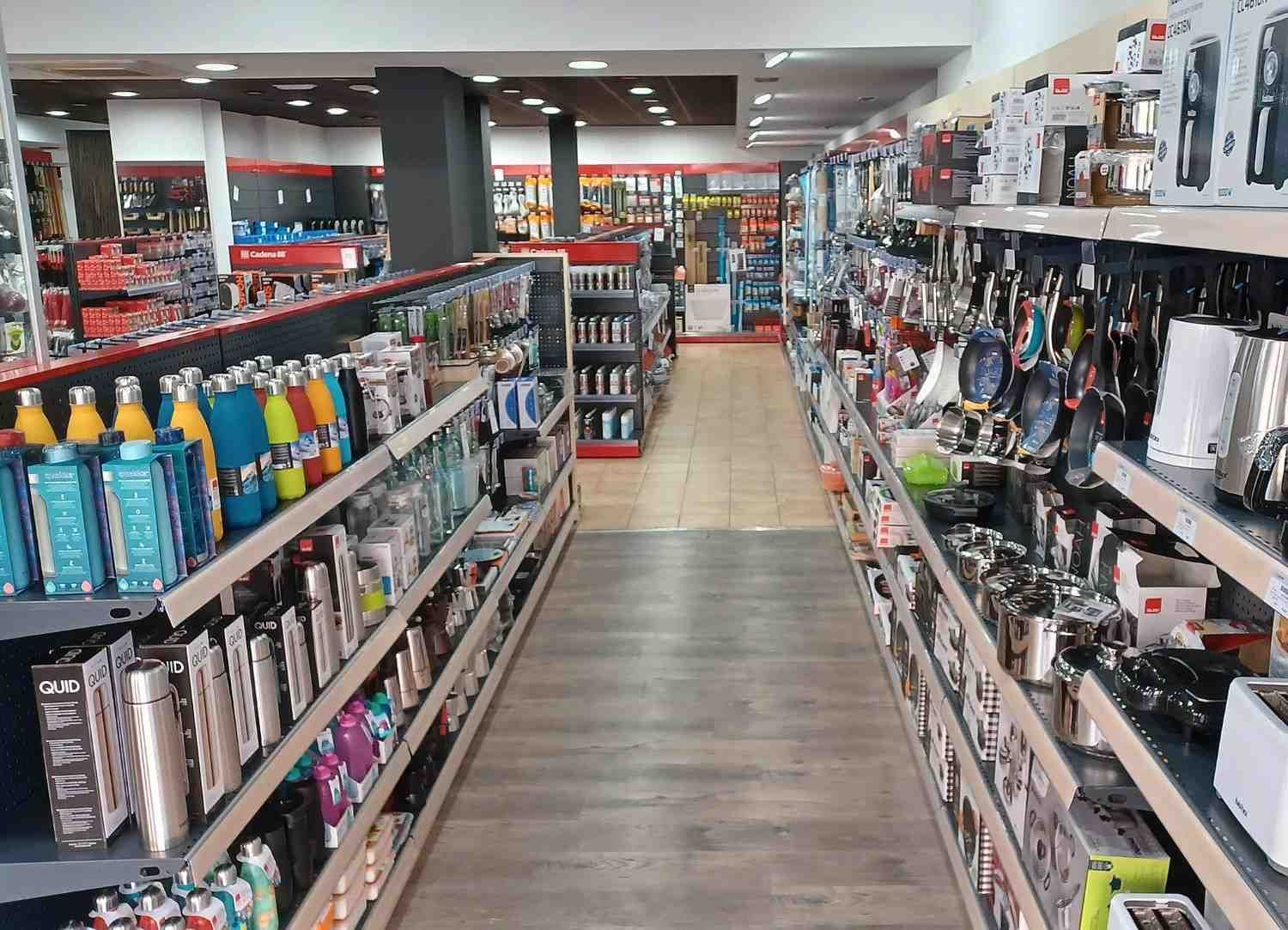 Vista interior de una tienda con filas de estantes repletos de diversos artículos para el hogar, muy iluminados.