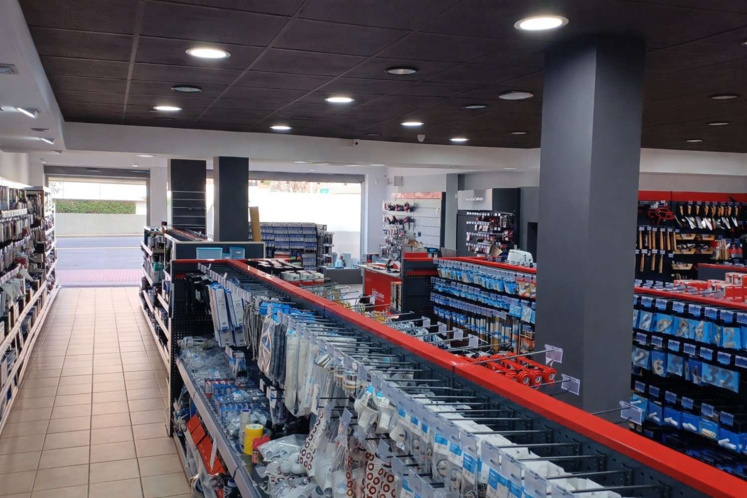 Interior de ferretería con pasillos de herramientas y suministros, detalles en rojo y un techo oscuro.