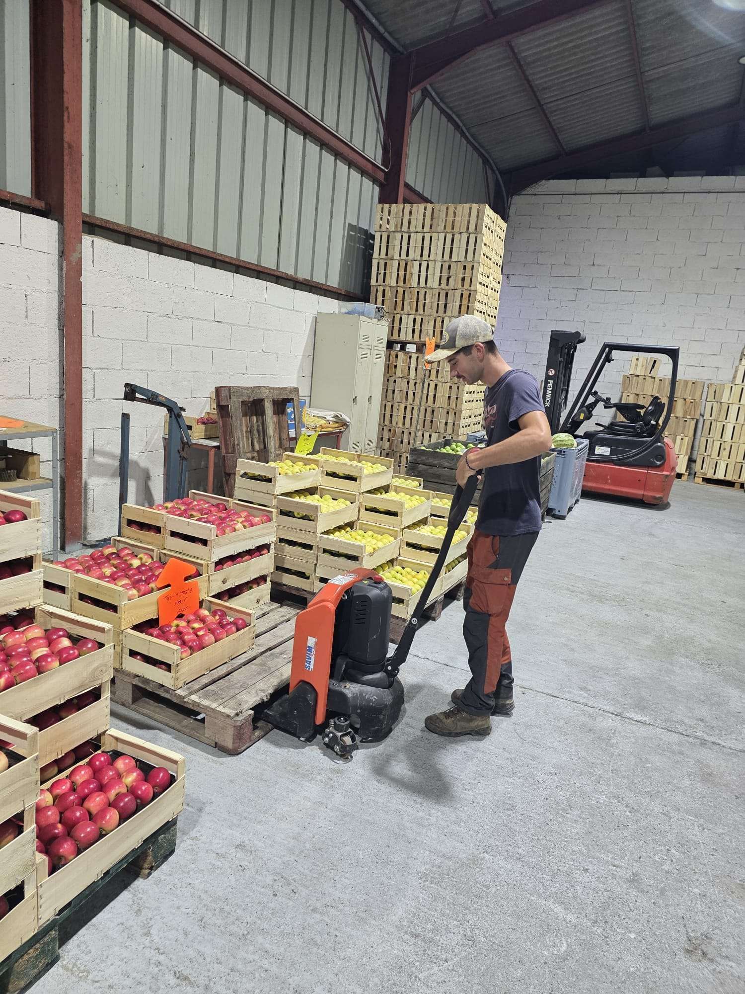 Un employé déplace un transpalette parmi des caisses empilées de produits rouges dans un entrepôt.