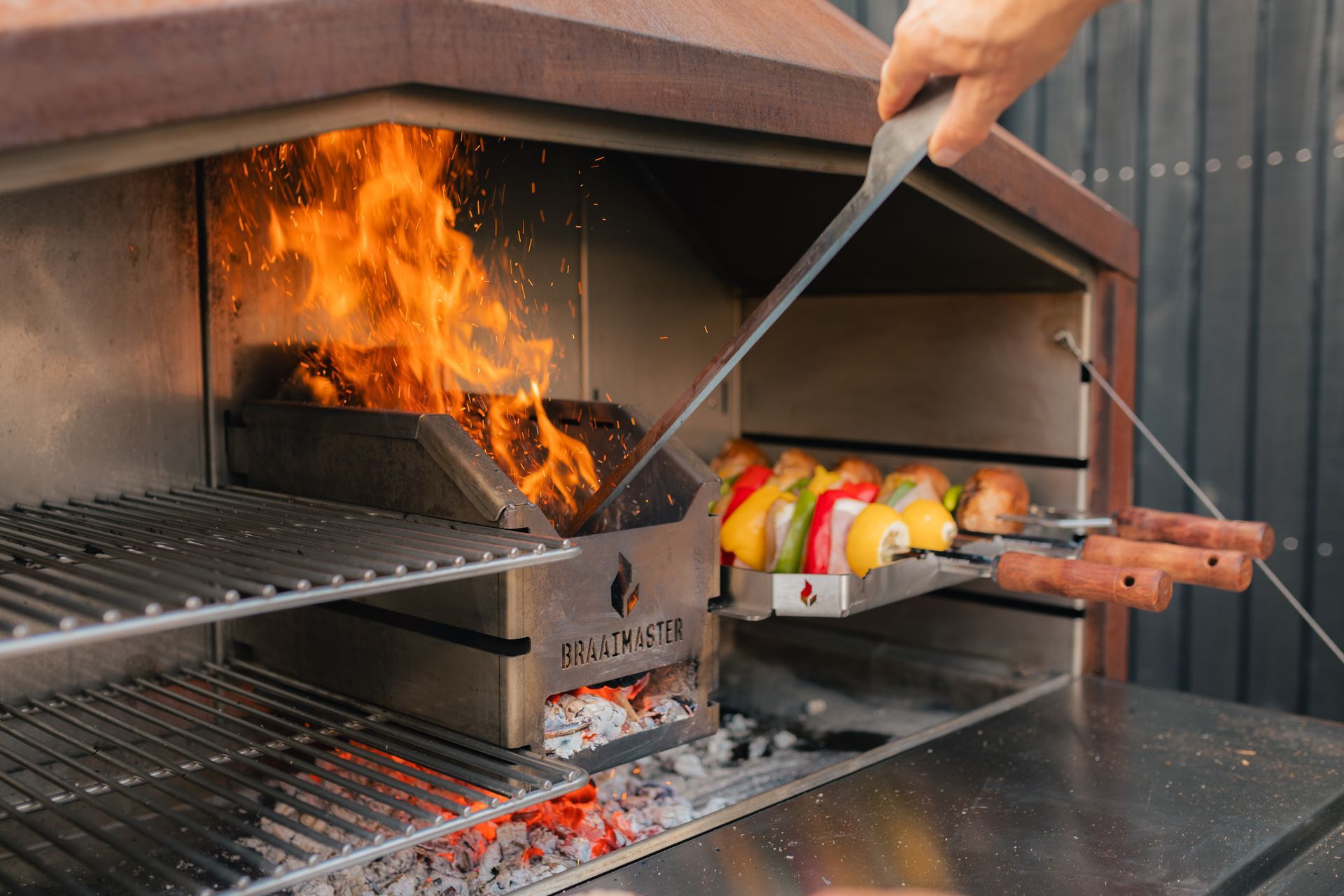 Braaimaster: Une personne fait griller des brochettes colorées sur les flammes d'un barbecue extérieur intégré.
