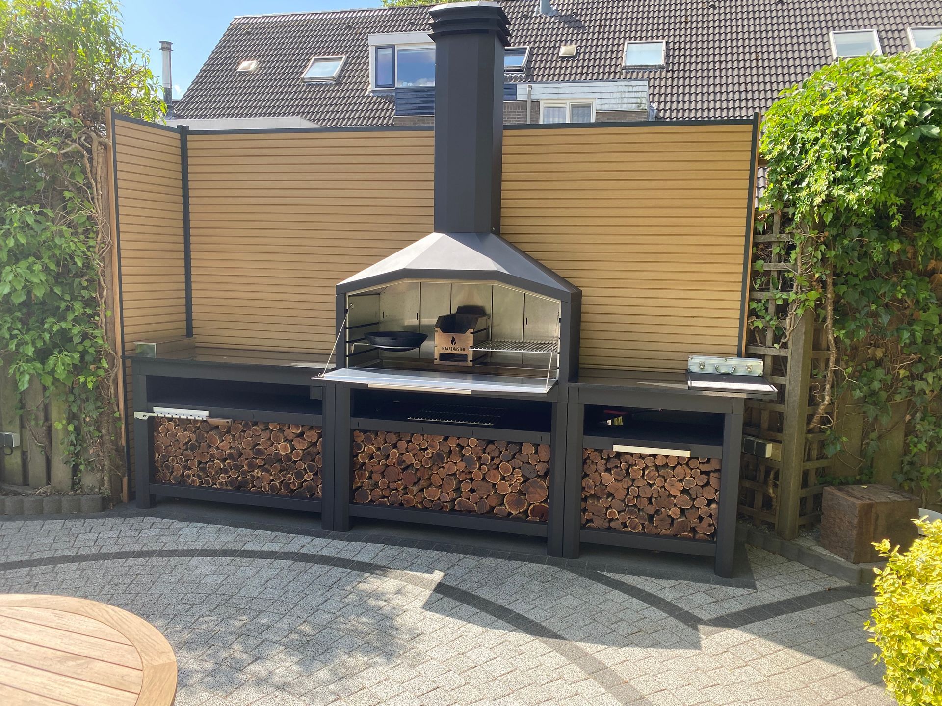 Braaimaster: Barbecue extérieur avec rangement pour le bois. Design noir et beige devant une clôture sur une terrasse.