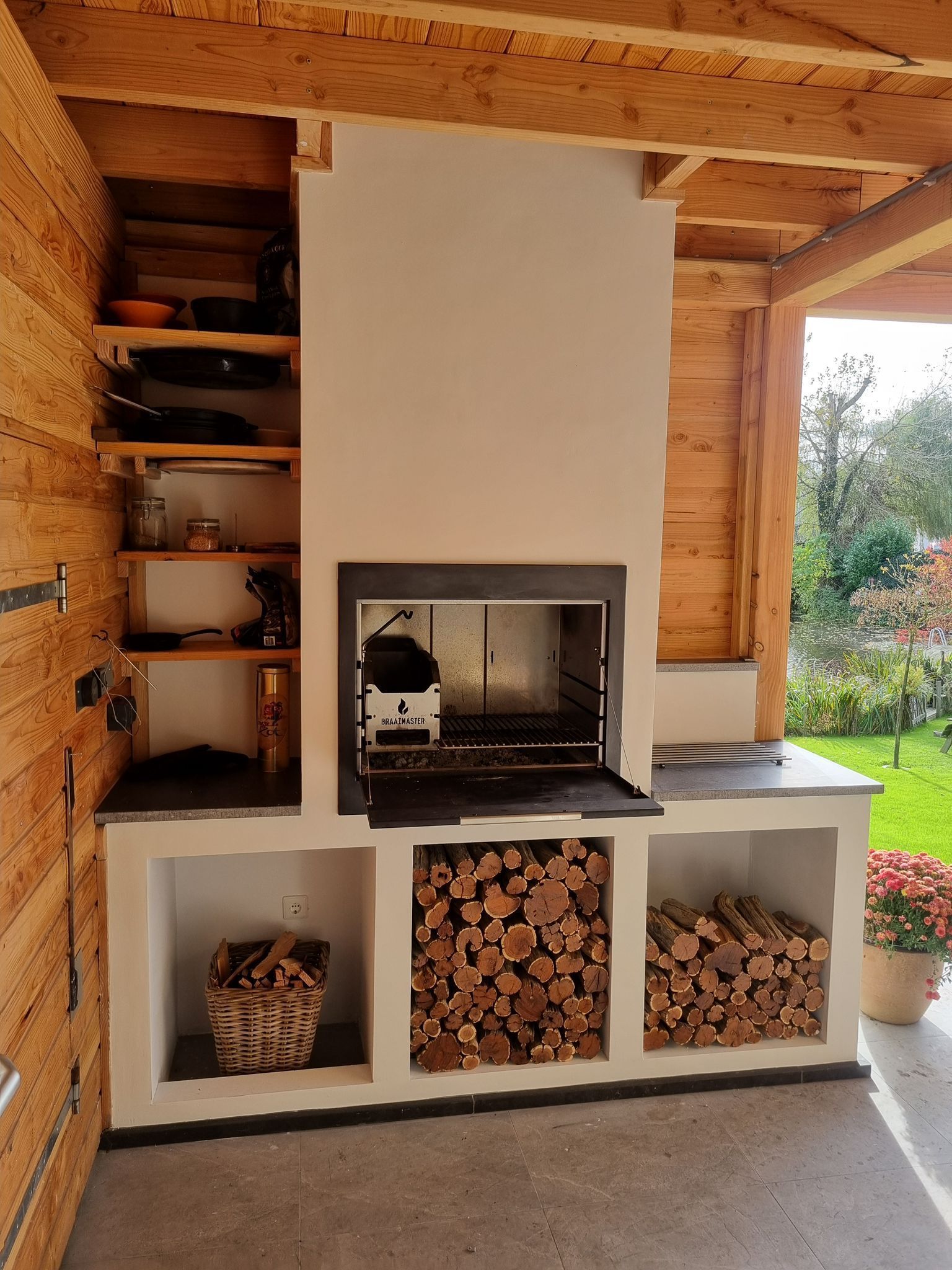 Braaimaster: Cuisine extérieure avec barbecue intégré, étagères et rangement pour le bois, sous un toit en bois.