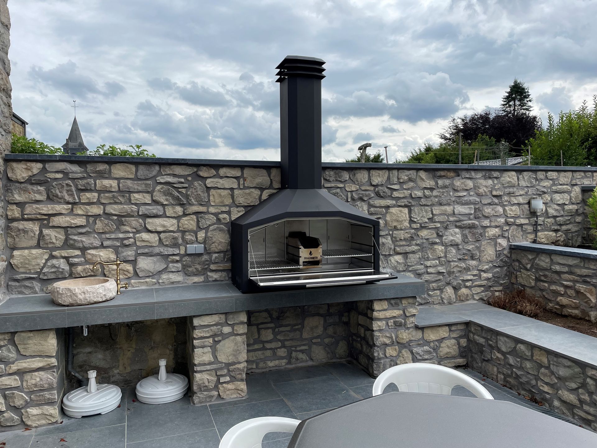 Braaimaster: Cheminée extérieure en pierre avec conduit et grille noirs, se détachant sur un ciel nuageux.