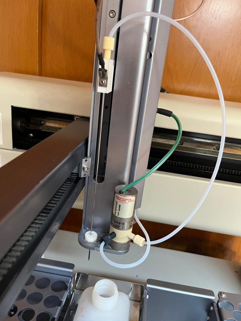 Primer plano del equipo de laboratorio: brazo robótico con tubos, conectado a una bomba y un vial de muestreo.