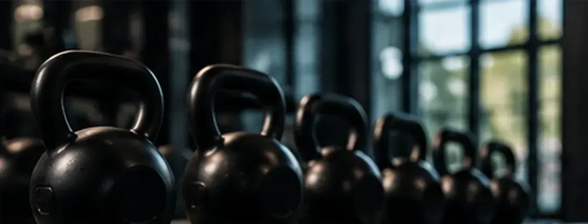 Una hilera de pesas rusas negras alineadas en un gimnasio con ventanas borrosas detrás.