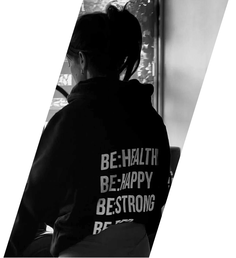 Una persona que viste una sudadera negra con capucha que dice “SÉ:SALUD, SÉ:FELIZ, SÉ:FUERTE” está sentada junto a una ventana.