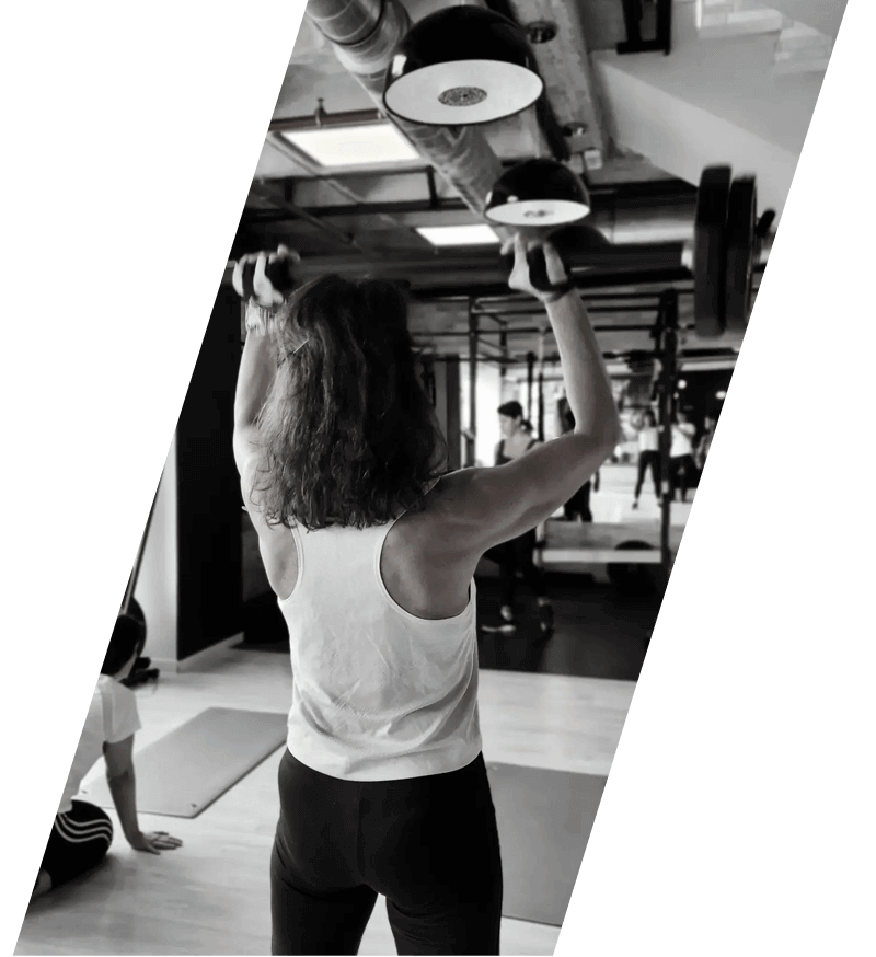 Una mujer en un gimnasio levanta mancuernas. Al fondo se ven otros usuarios y equipos.