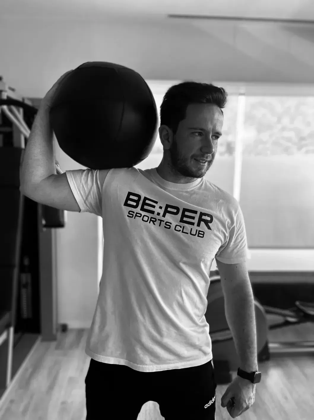 Un hombre sostiene una gran pelota negra de ejercicio al hombro en un gimnasio. Lleva una camiseta blanca y pantalones negros.