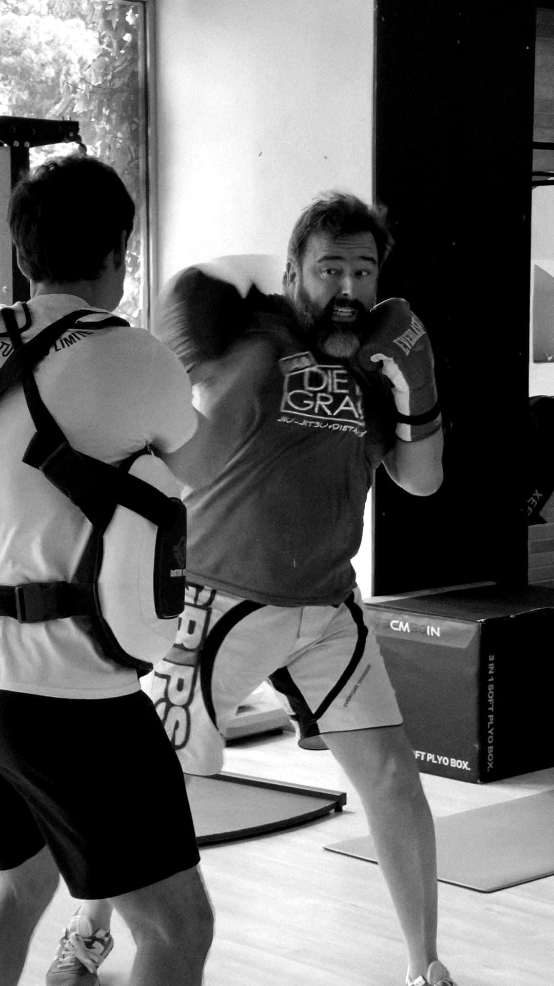 Una persona vestida con indumentaria de boxeo golpea un saco de entrenamiento que sostiene un entrenador dentro de un gimnasio.
