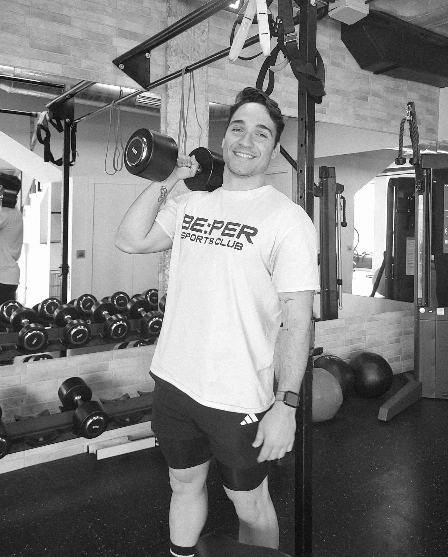 Una persona sonriente, vestida con una camiseta deportiva, se encuentra en un gimnasio sosteniendo una mancuerna a la altura de los hombros.