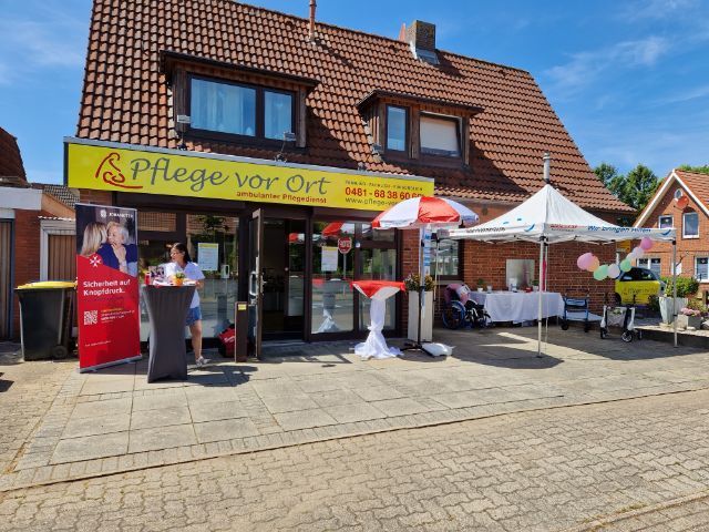 Straßencafé mit rot-weißen Sonnenschirmen und einem gelben „Pflege vor Ort“-Schild an einem Backsteingebäude
