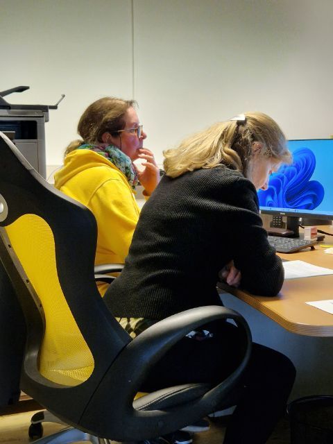 Zwei Personen sitzen an Schreibtischen in einem Büro und benutzen Computer; eine trägt einen gelben Kapuzenpullover, die andere eine schwarze Jacke.