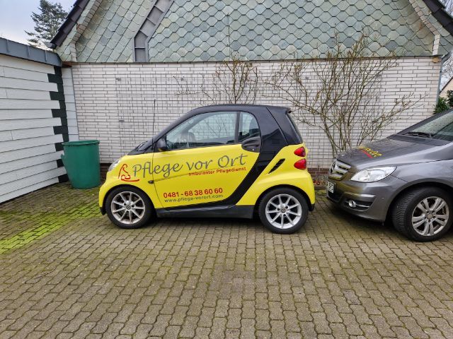 Ein gelb-schwarzer Smart mit der Aufschrift „Pflege vor Ort“ parkt neben einem grauen Auto in einer Einfahrt.
