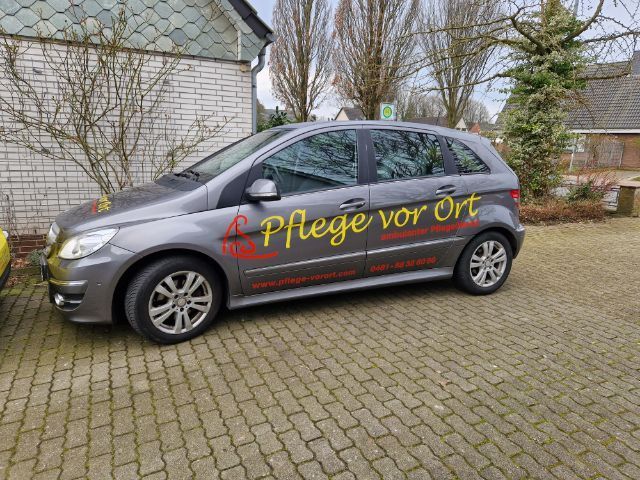 Grauer Lieferwagen mit der Aufschrift „Pflege vor Ort“ parkt auf einer gepflasterten Auffahrt.