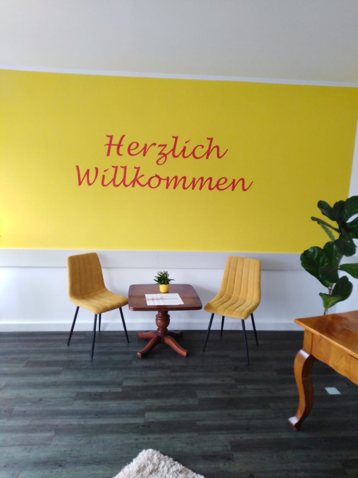 Gelbe Wand mit der Aufschrift „Herzlich Willkommen“, zwei senffarbene Stühle, ein kleiner Tisch und eine Topfpflanze in einem hellen Raum