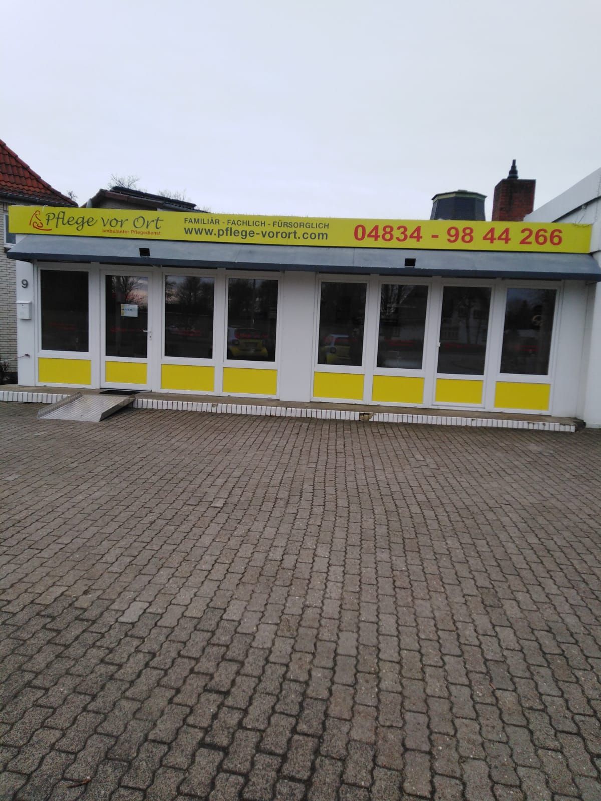 Gelbe Ladenfront mit großen Fenstern und einem Schild, von einer Kopfsteinpflasterstraße aus gesehen.