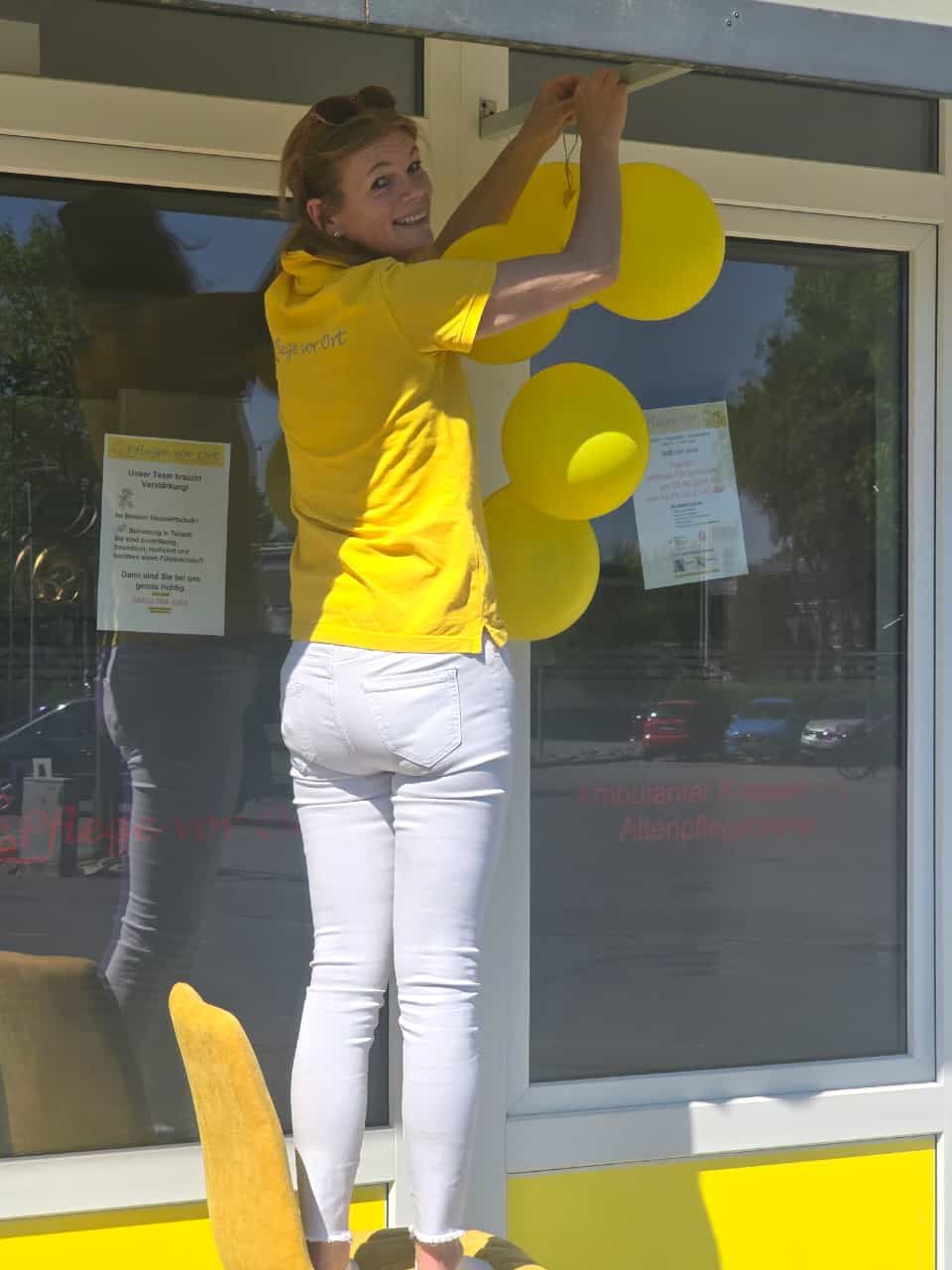 Eine Person in einem gelben Hemd und einer weißen Hose hängt gelbe Luftballons an ein Schaufenster.